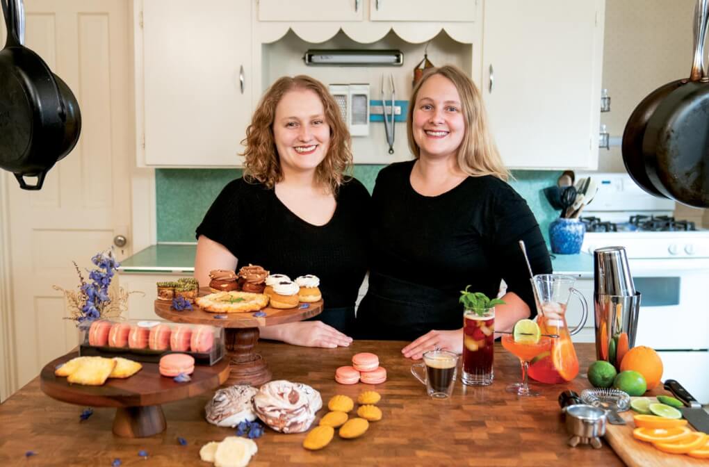 Two sisters stand in a kitchen surrounded by colorful pastries and drinks